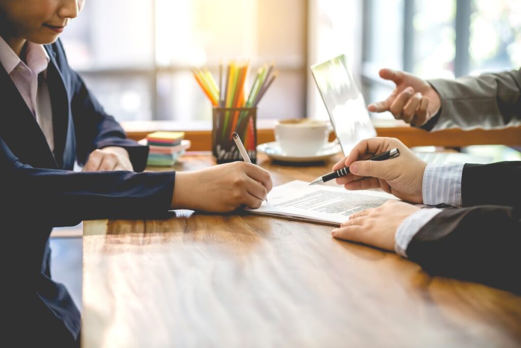 Foto com 3 mãos em cima de uma mesa assinando contratos e explicando termos