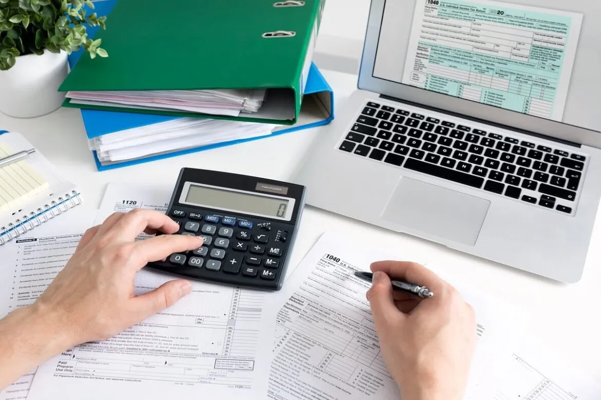 Pessoa em uma mesa com notebook, documentos e uma calculadora
