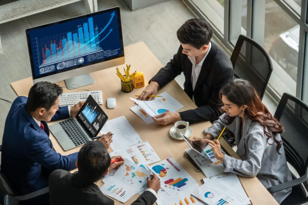 Mesa de trabalho com ao menos quatro pessoas, analisando gráfico em computador