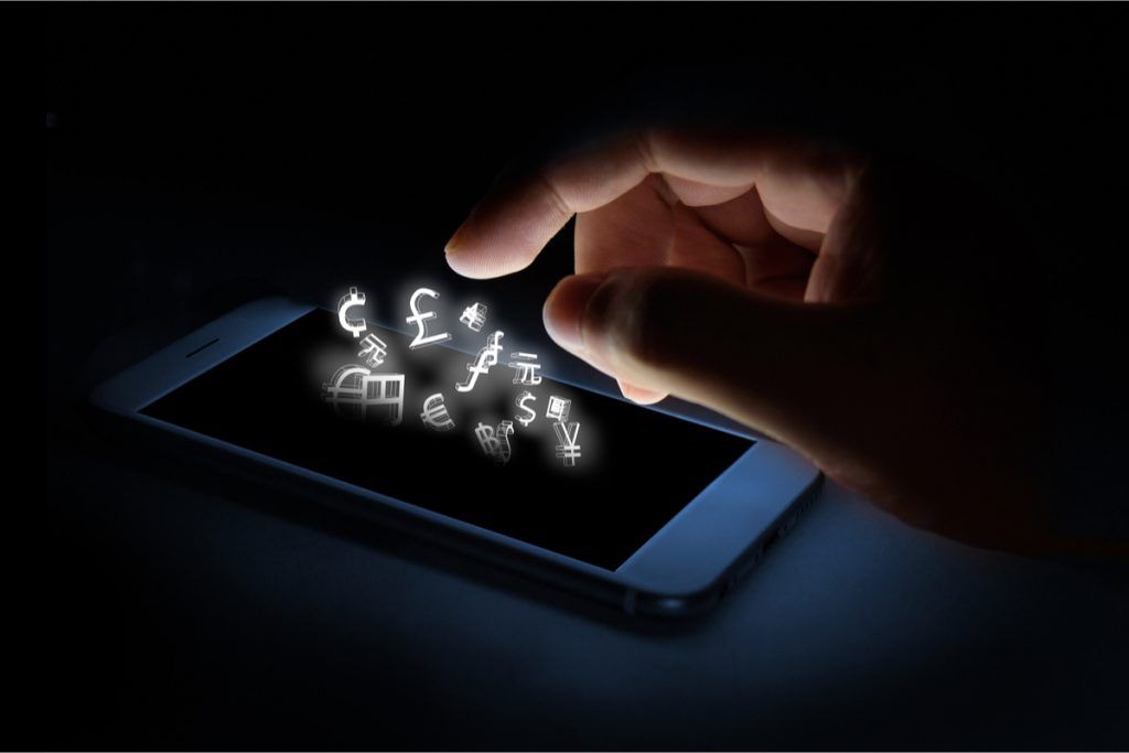 Imagem escura apenas iluminada por ícones financeiros que saem da tela de um smartphone tocado por uma mão humana.