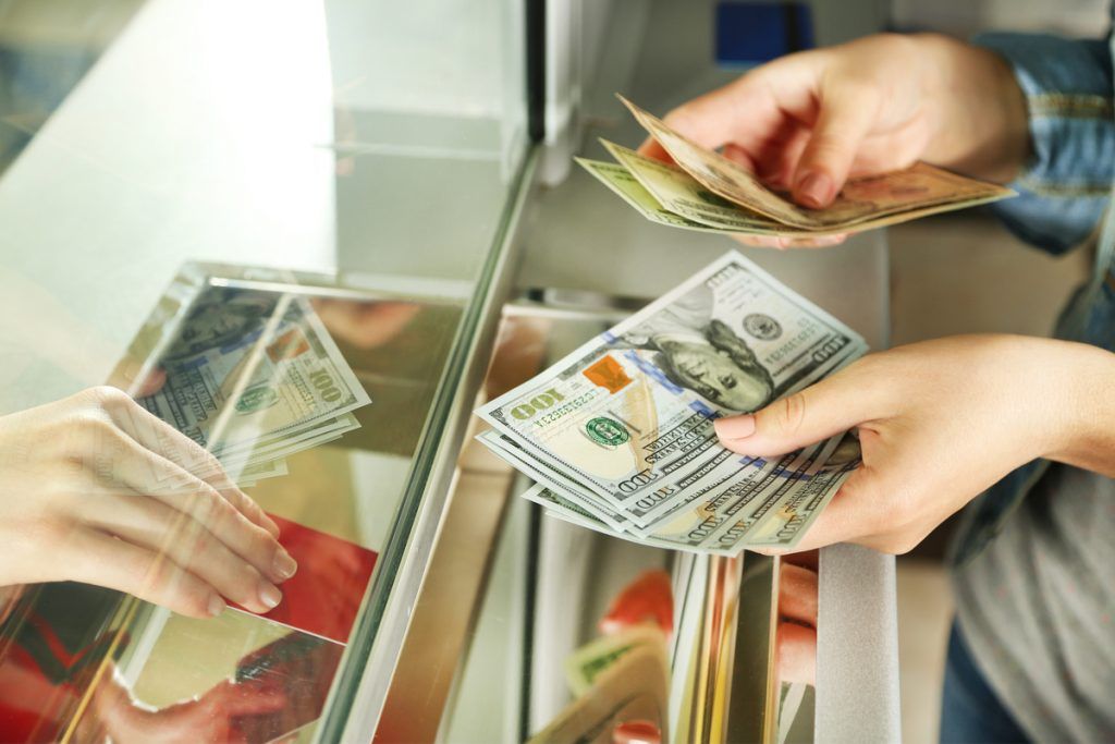 Imagem de uma pessoa contando notas de moedas de diferentes países, com close nas mãos da pessoa por trás de uma vidraça, como se estivesse em um caixa de casa de câmbio