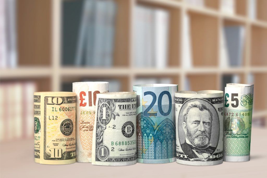 Imagem de notas de dinheiro de países diversos, enroladas e empilhadas de pé sobre uma mesa.
