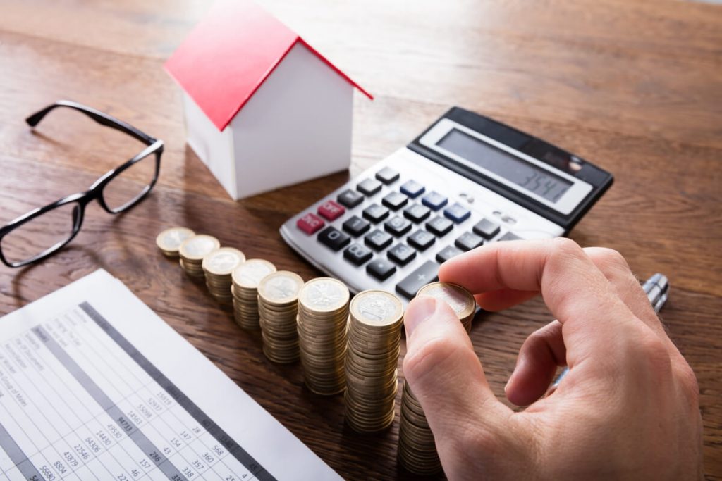 Uma mesa com objetos como um óculos, papíes, calculadora e a miniatura de uma casinha, com a mão de uma pessoa empilhando moedas sequencialmente