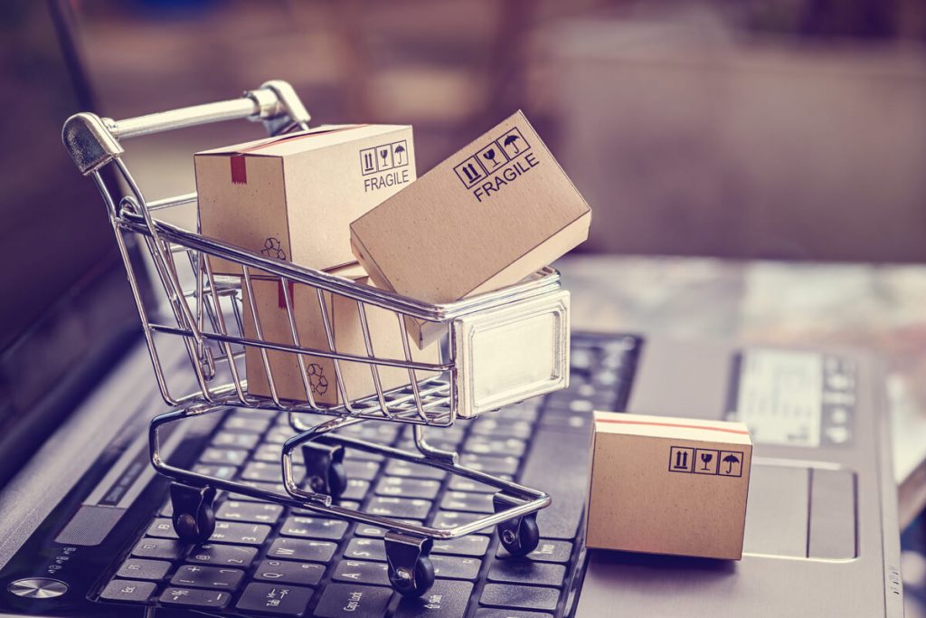 Miniatura de um carrinho de supermercado cheio de caixas de papelão e sobre um notebook para ilustrar um e-commerce