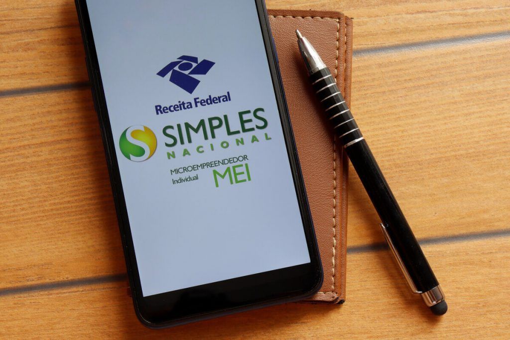 Imagem de um smartphone que exibe na tela o logotipo do Simples Nacional, da Receita Federal. Ao lado uma agenda e uma caneta.