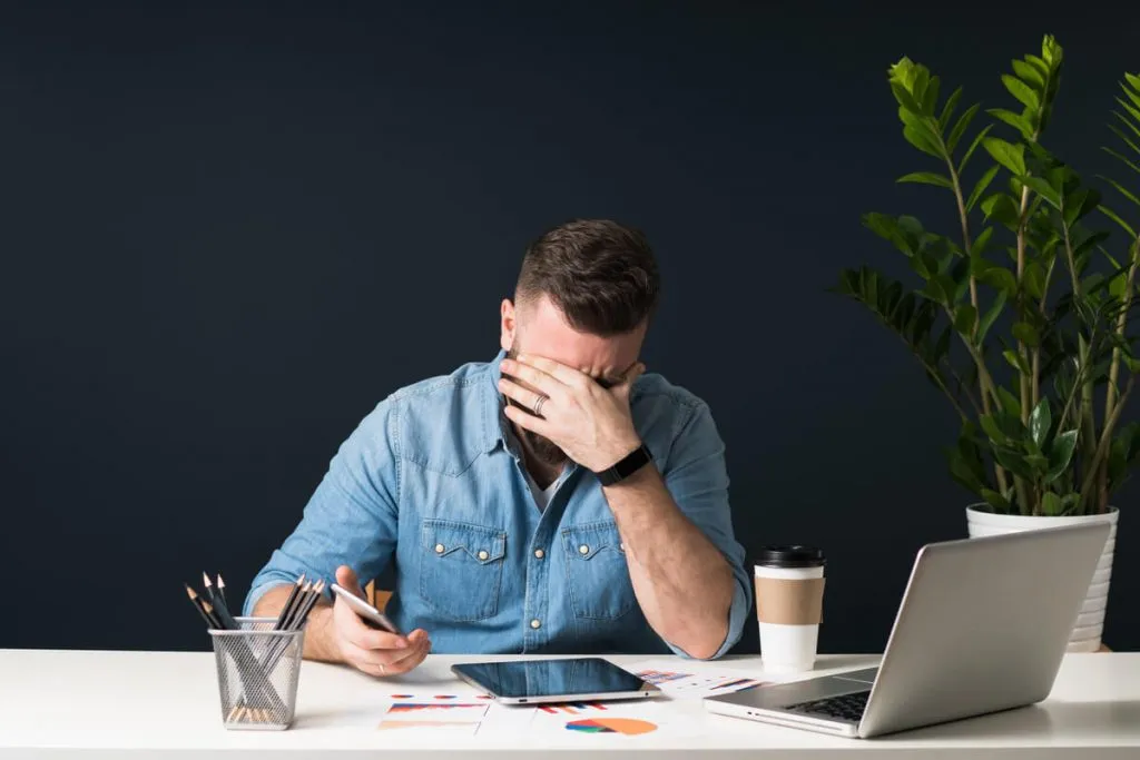 Homem em uma mesa com a mão sobre o rosto aparentando cansaço. Na outra mão um notebook, à sua frente um tablet, um notebook, um copo de café, um porta lápis e documentos espalhados.