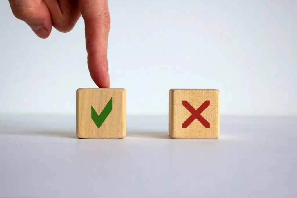 Imagem de duas peças de madeira pequenas, uma com X vermelho e a outra com um V verde, tocado por um dedo humano.