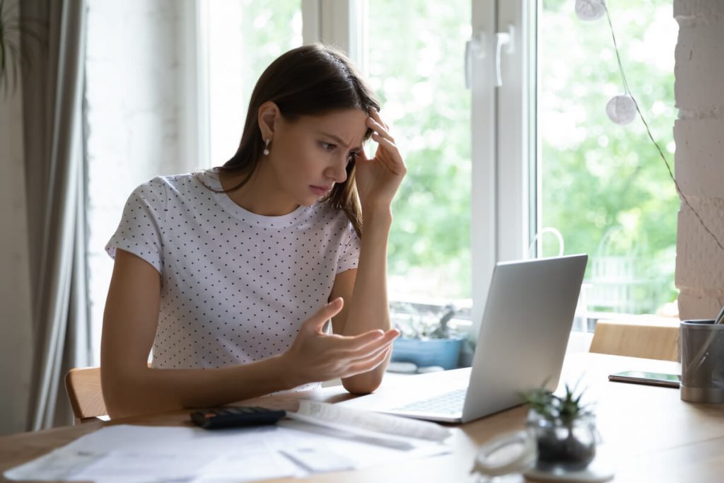 Jovem moça de camiseta branca sentada em frente ao seu notebook e com expressão de preocupação e incredulidade no rosto, além e vários papeis espalhados pela mesa, representando despesas