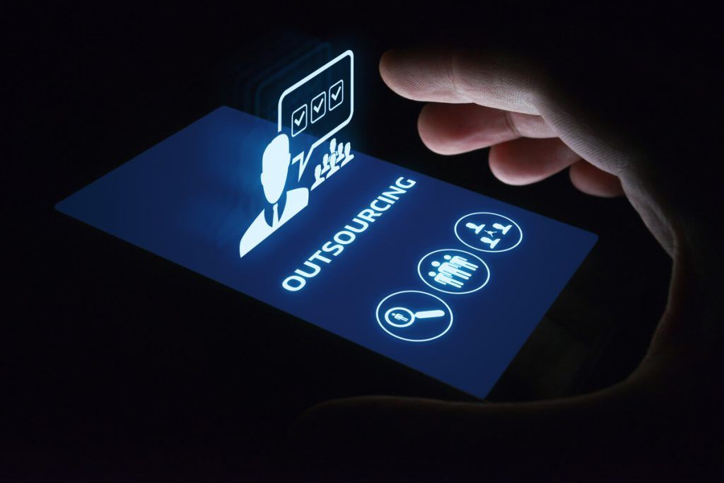 Uma mão humana tocando um smartphone no escuro, onde se lê a palavra outsorcing com vários icones na tela representando comunicação