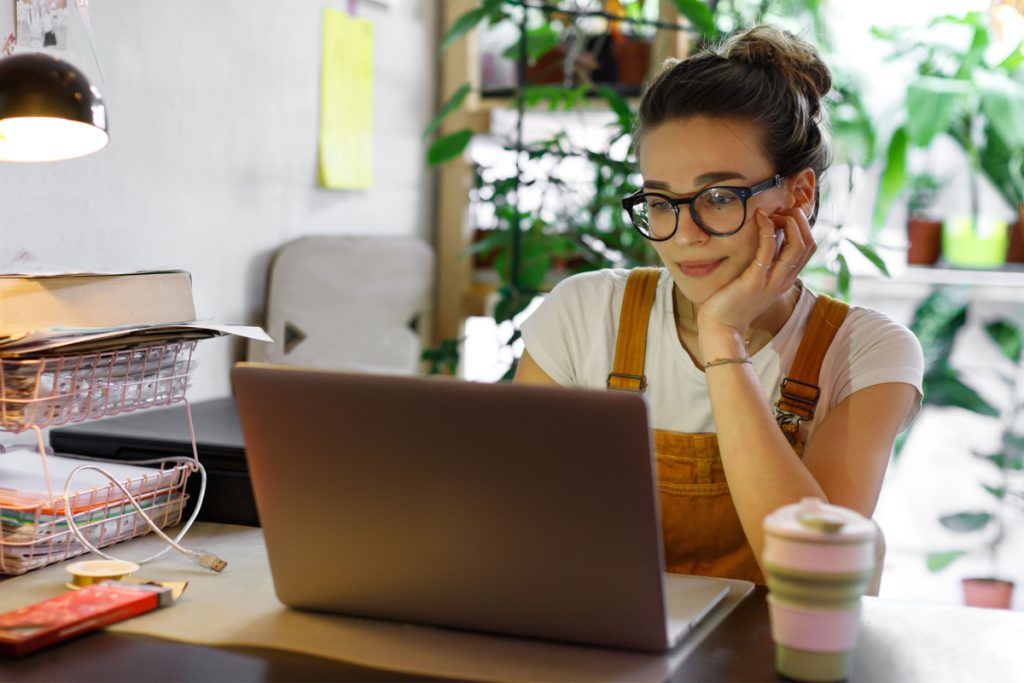 Moça jovem e com semblante sereno, sentada em uma mesa de trabalho com um copo de café especial, utilizando seu notebook