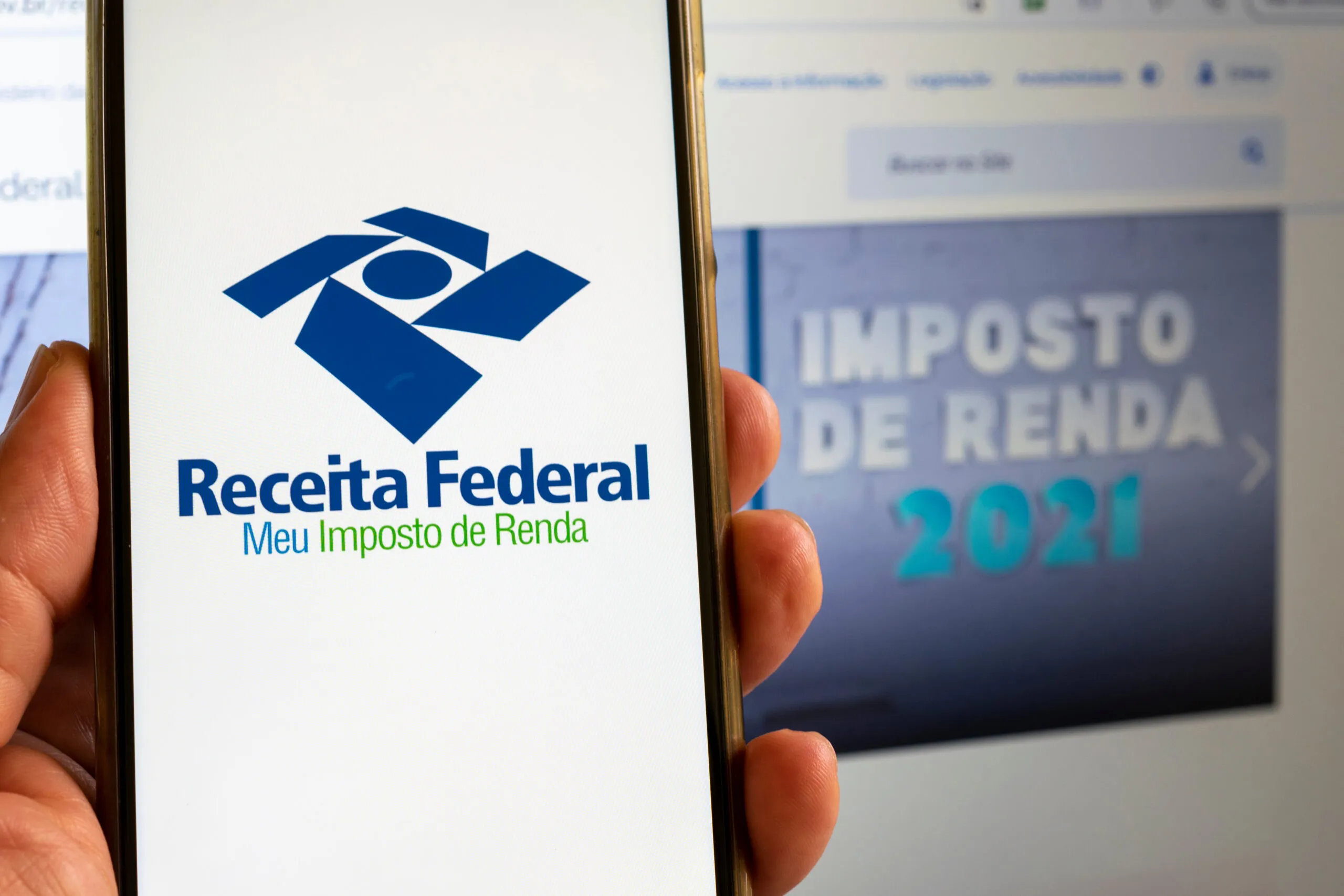 Imagem de um celular acessando o site da Receita Federal e ao fundo um computador com uma imagem destacada onde se lê Imposto de Renda 2021