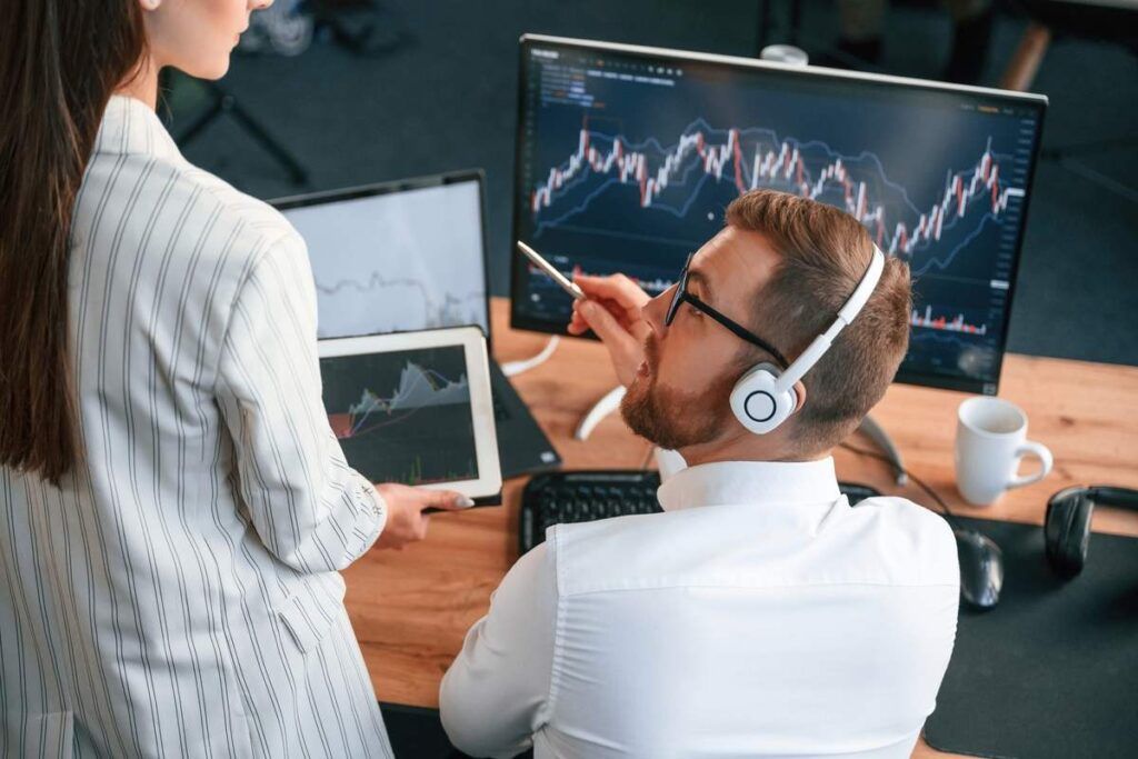Investidor fazendo especulação financeira com colega de trabalho e computador com desempenho financeiro ao fundo. 