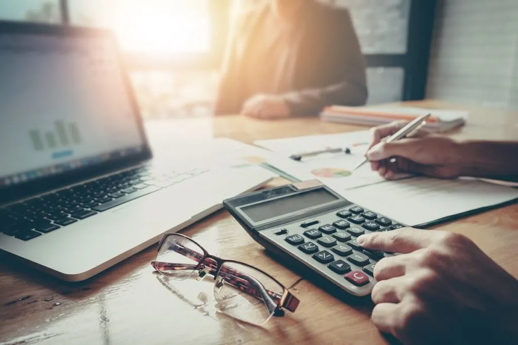 Pessoa em uma mesa com calculadora, um óculos ao lado e um notebook aberto