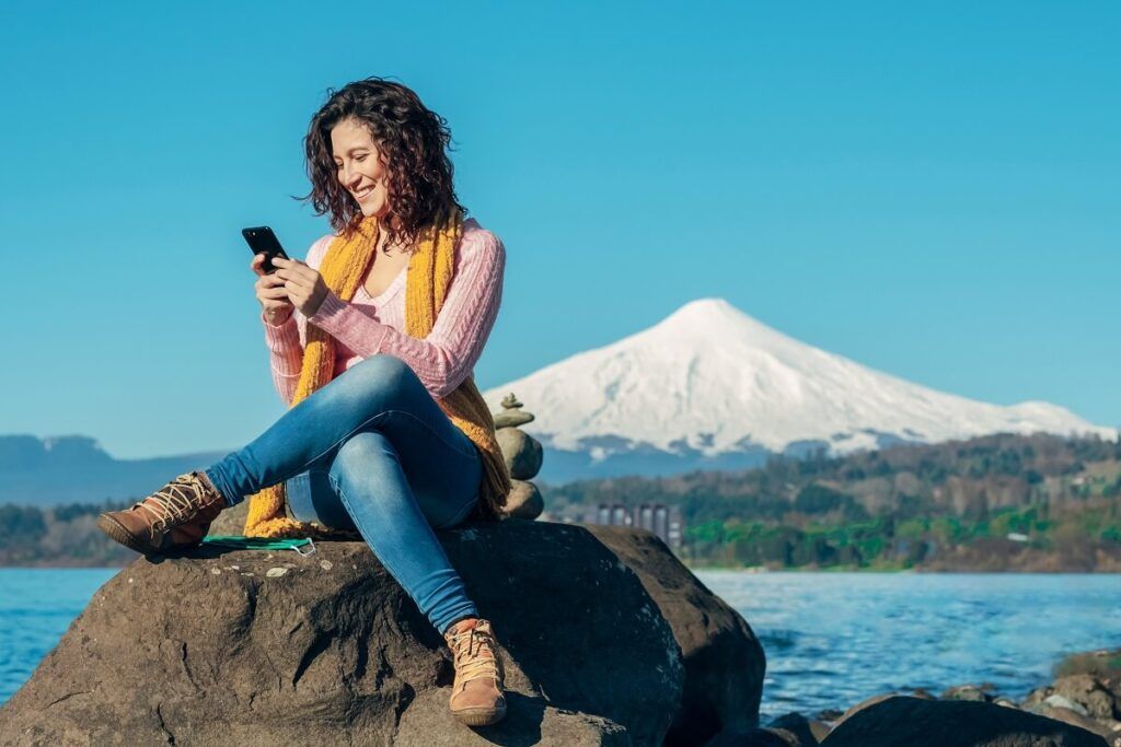 Mulher usando o celular em cima de uma pedra e ao fundo uma linda paisagem de uma montanha com gelo