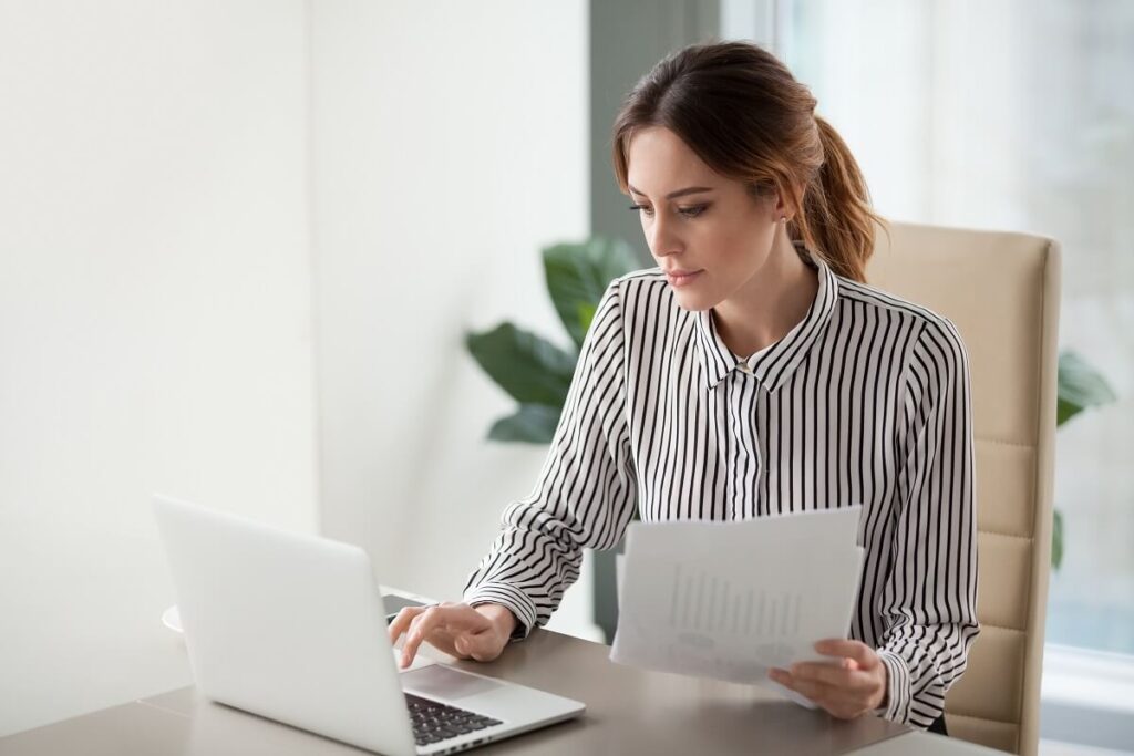 Mulher com um documento na mão e com a outra mexendo no notebook