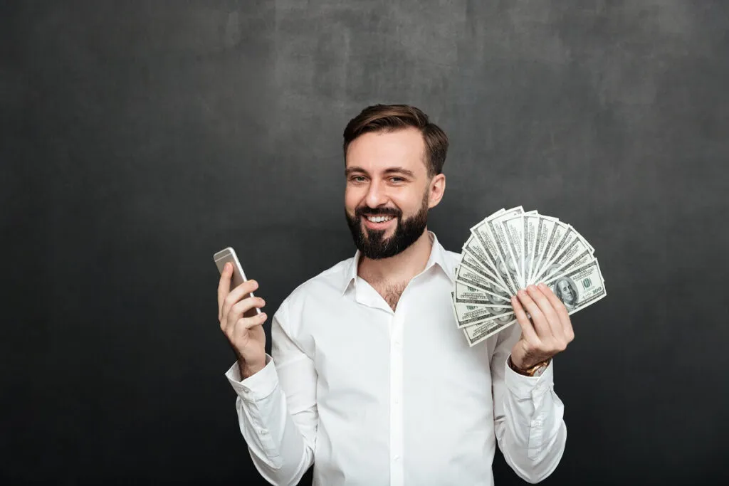Homem de camisa social branca segurando notas de dólar em uma mão e na outra o celular