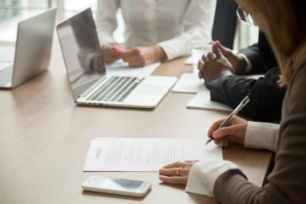 Pessoa assinando uma proposta comercial Uma pessoa assinando uma proposta comercial, acompanhada de outras duas em uma mesa de reunião.