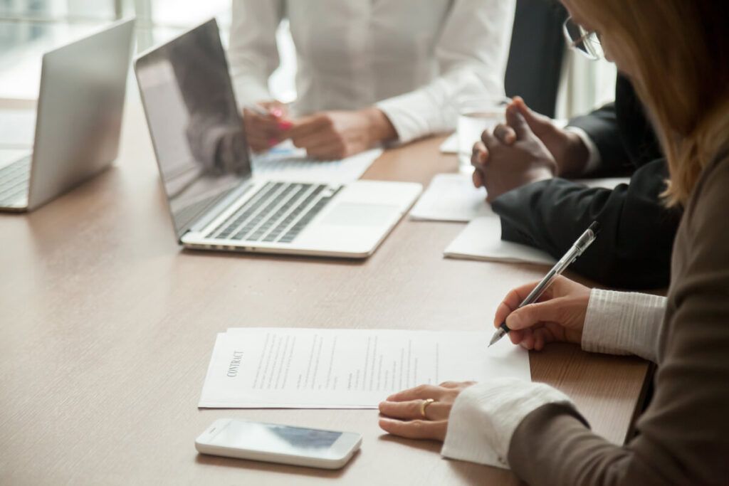 Uma pessoa assinando uma proposta comercial, acompanhada de outras duas em uma mesa de reunião.