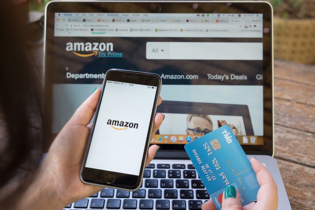 Mulher que pretende vender online, de frente para tela de um notebook com a página da Amazon aberta, um smartphone em uma das mãos e o cartão de crédito em outra.