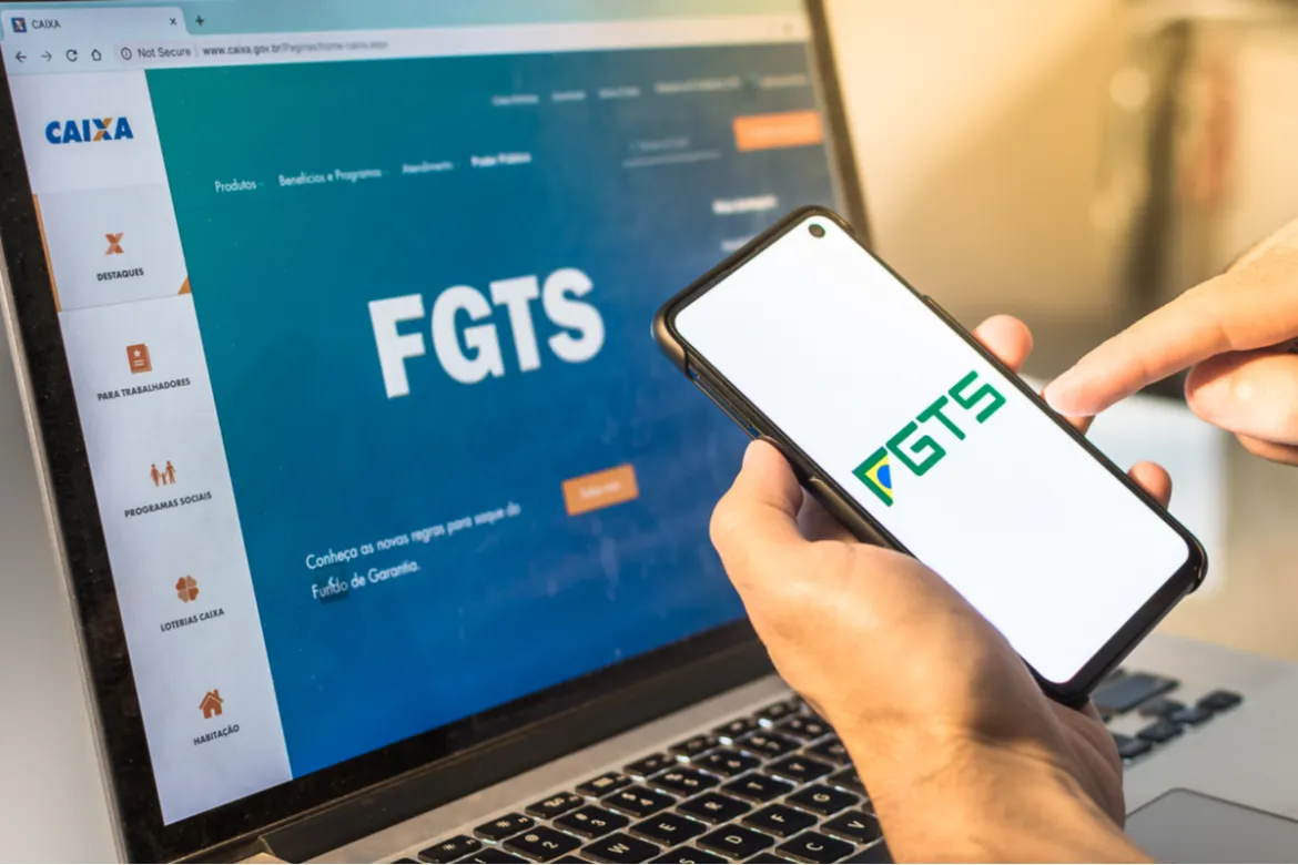 Pessoa segurando um celular com o logo do FGTS da tela, em segundo plano, um notebook com o site do FGTS aberto