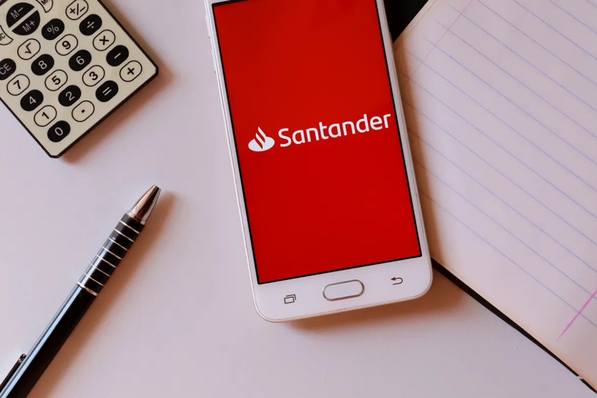 celular com o aplicativo aberto do Santander ao lado de um caderno, caneta e calculadora em cima da mesa