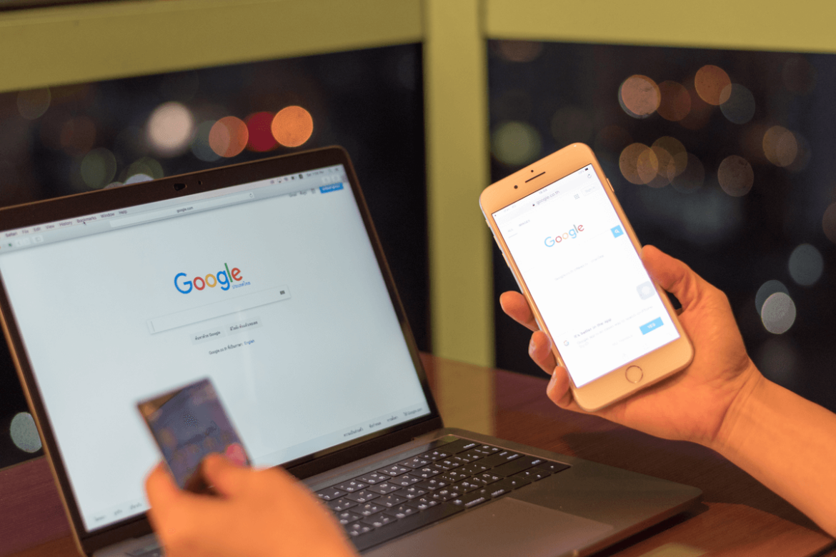 Imagem de uma mão segurando um celular com a página do google aberta, um cartão na outra mão em frente a um notebook que tem a tela do Google aberta