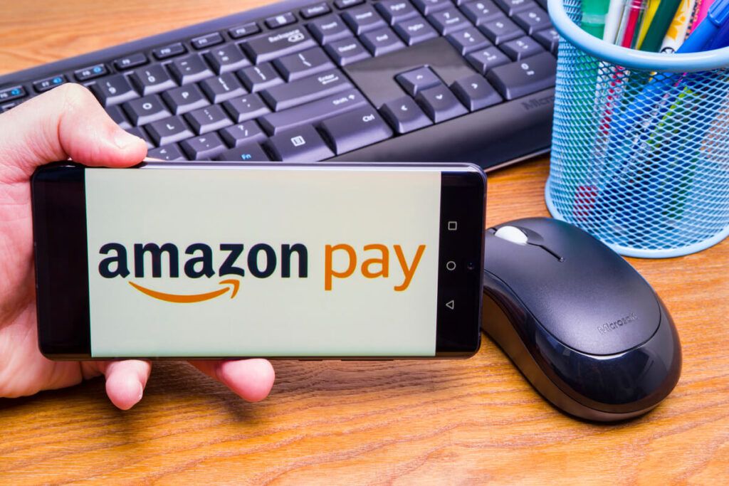 Mesa com teclado e mouse, e uma mão segurando um smartphone com a tela da Amazon Pay.