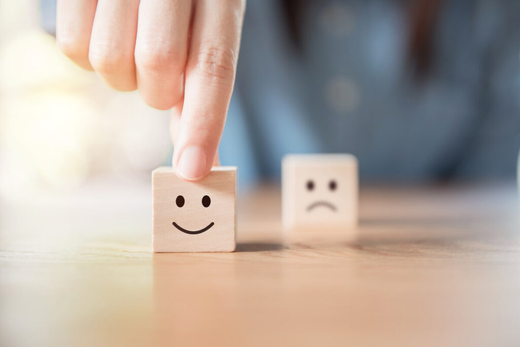 Dois pequenos cubos de madeira representando o que é feedback. Um com um rostinho feliz, em destaque, outro com um rostinho triste, desfocado na imagem.