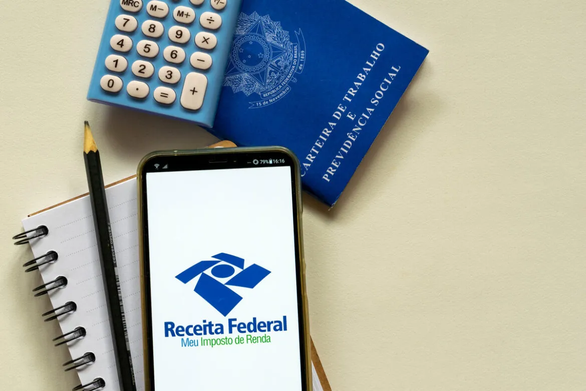 Uma mesa vista de cima com um celular acessando o site da Receita Federal, um carteira de trabalho e uma calculadora