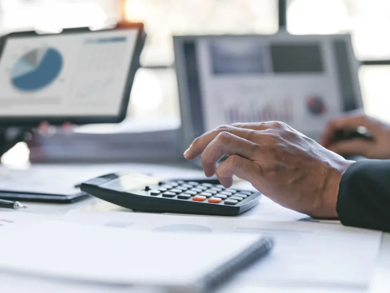 Mãos de um profissional utilizando uma calculadora sobre documentos, com telas de gráficos ao fundo, ilustrando a gestão do Imposto de renda MEI.
