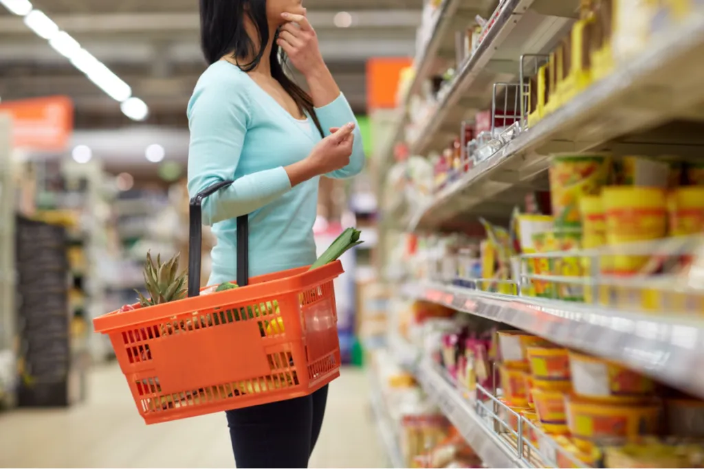 Mulher em corredor de supermercado segurando uma cesta de compras, olhando para uma prateleira e pensando