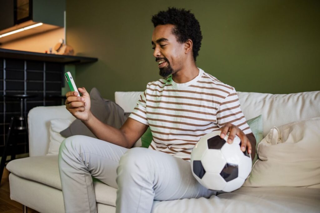 Homem sentado no sofá acompanhando o jogo pelo celular.