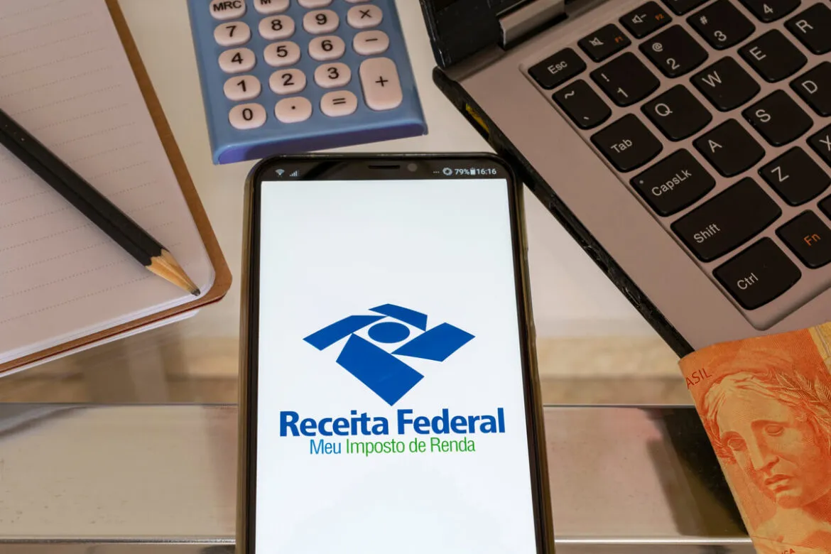 Celular com o aplicativo da Receita Federal na tela, em cima de uma mesa ao lado de uma calculadora, computador, caderno e lápis