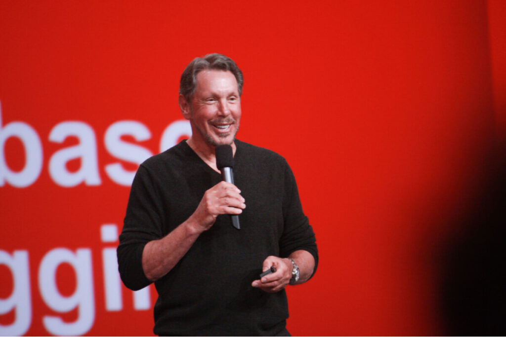 Foto de Larry Ellison segurando um microfone em frente à um painel vermelho