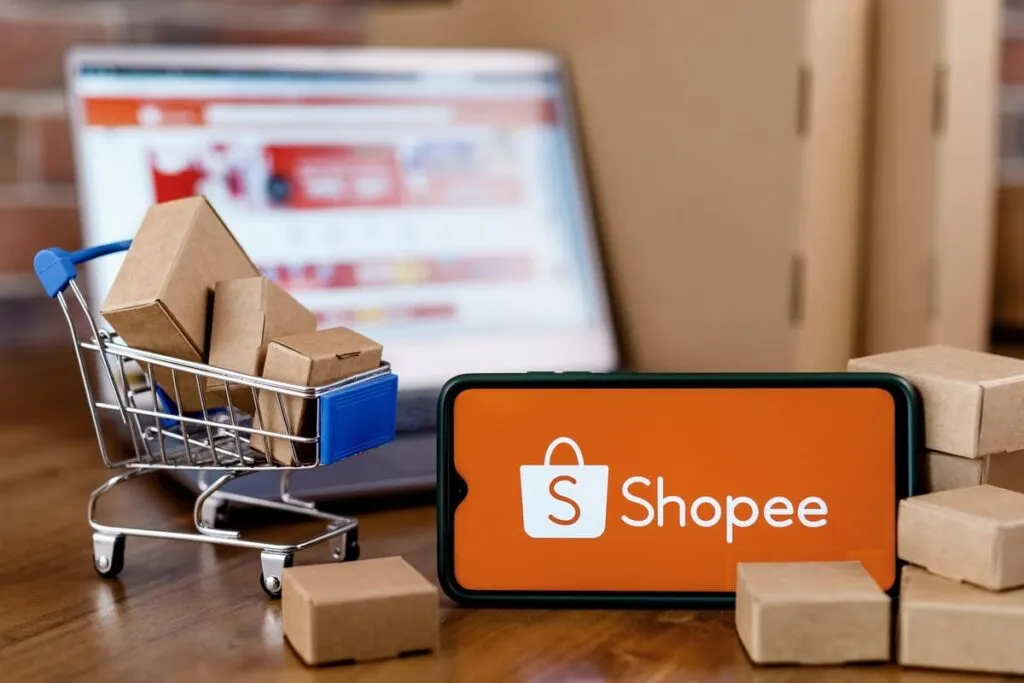 Aplicativo da Shopee aberto em um celular ao lado de um carrinho de compras em miniatura.