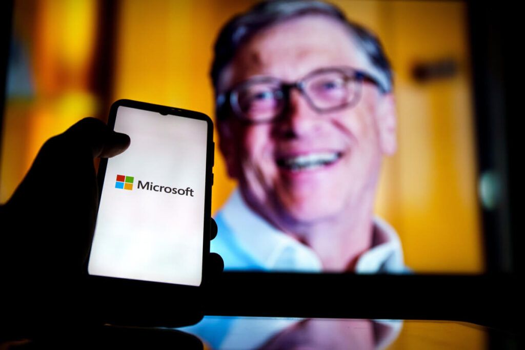 Foto de um celular com o logotipo da empresa Microsoft e, ao fundo, imagem de Bill Gates