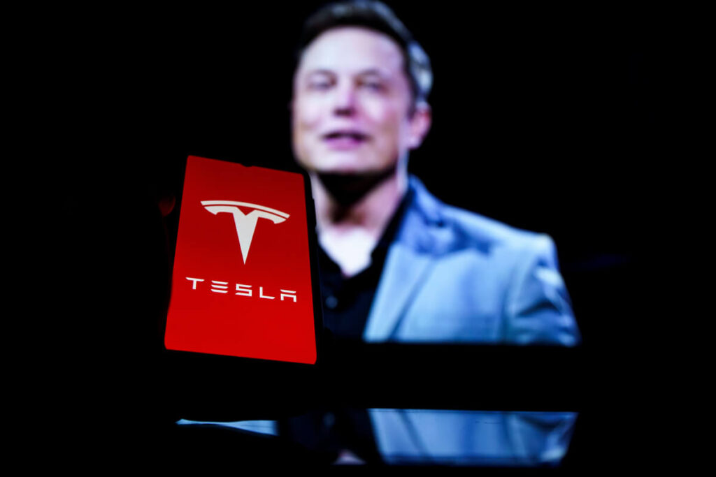 Imagem de um celular com o logo da empresa Tesla e, ao fundo, e Elon Muk