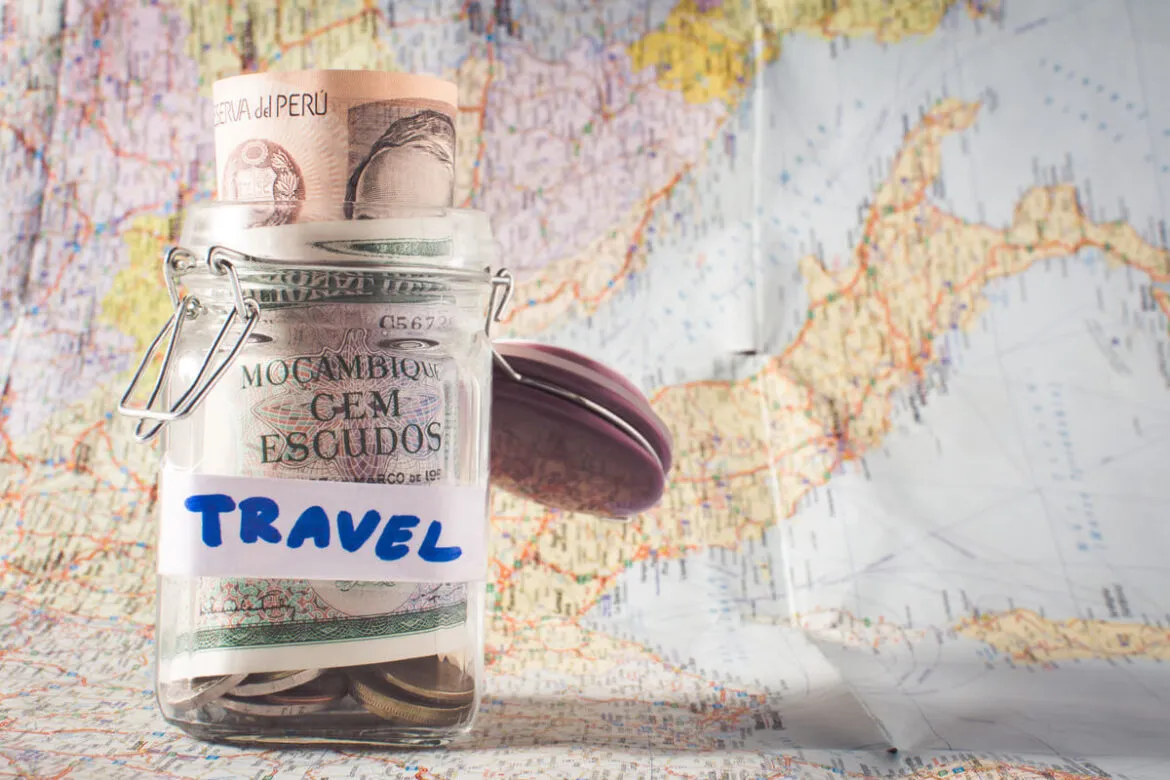 pote com etiqueta escrito "travel" cheio de dólares e, em segundo plano, um mapa mundi