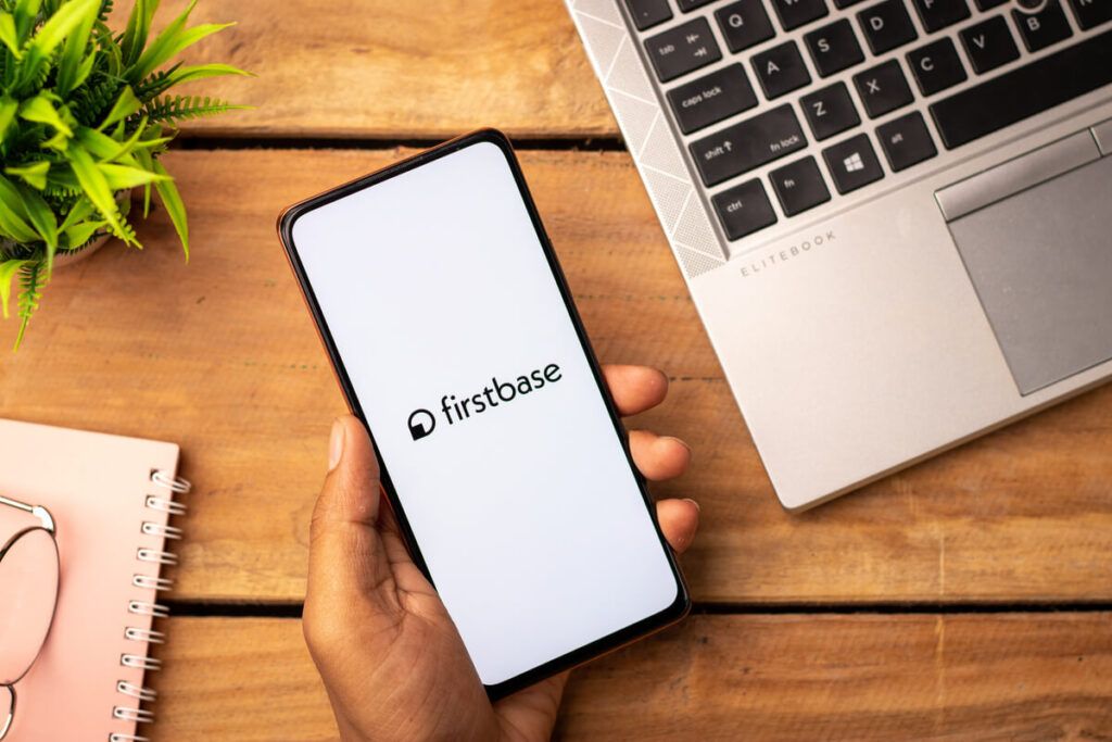 Uma visão de cima de uma pessoa segurando um smartphone com o logo da Firstbase aparecendo na tela.