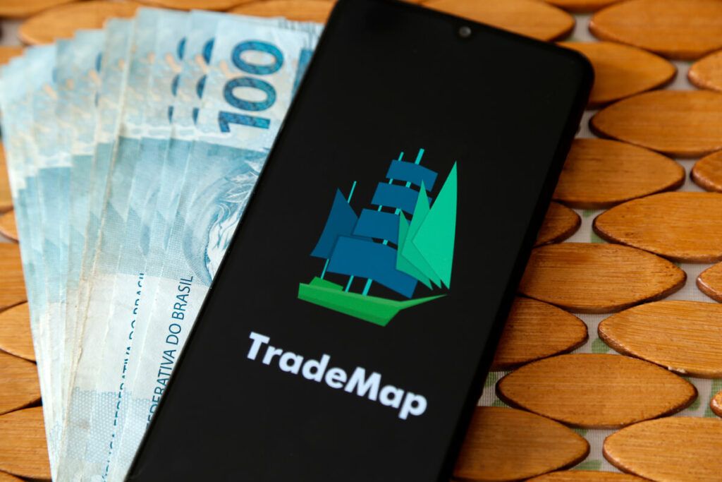 Imagem de várias notas de 100 reais e sobre elas um celular com o logo da TradeMap