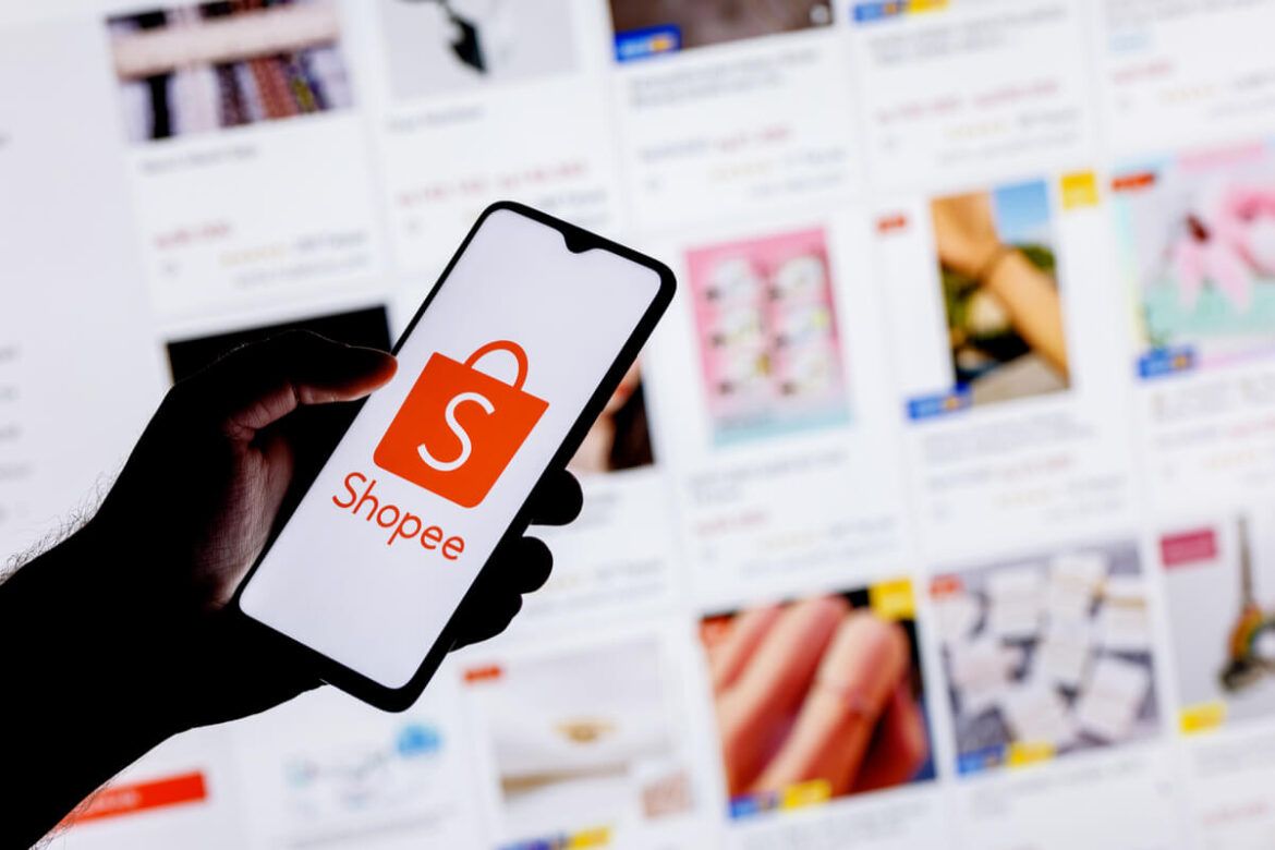 Uma pessoa segurando um celular com o logo da Shopee na tela e ao fundo várias imagens de produtos em uma tela
