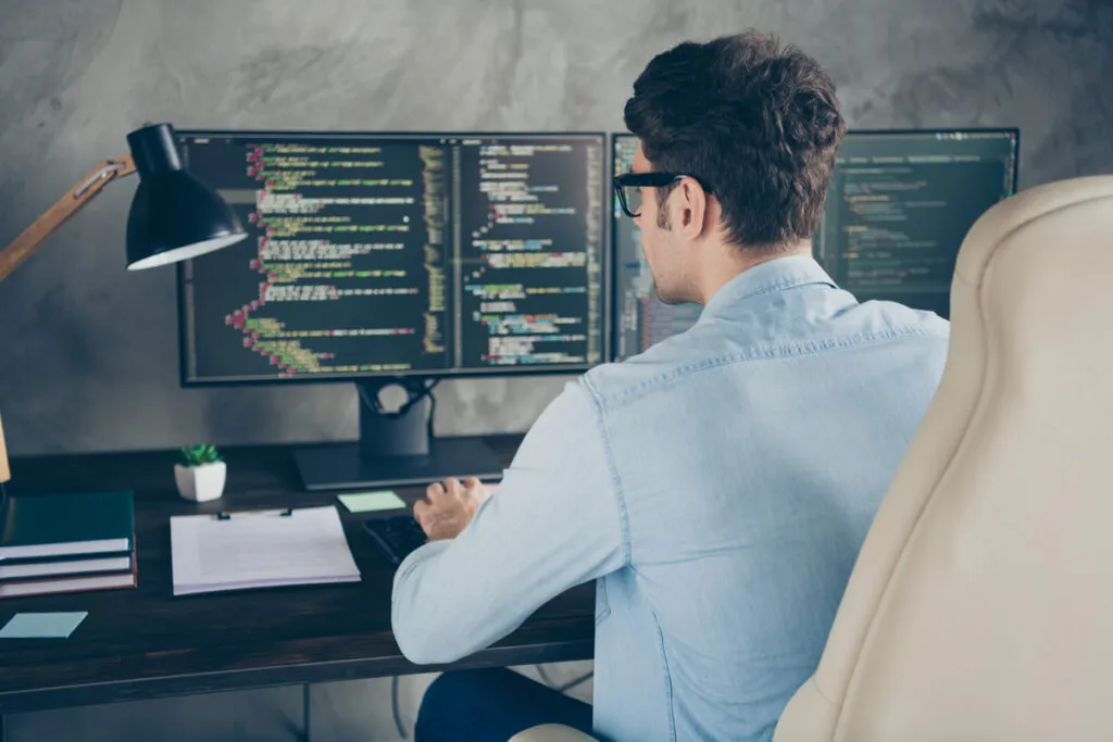 Homem sentado em frente a um computador com códigos de programação abertos na tela