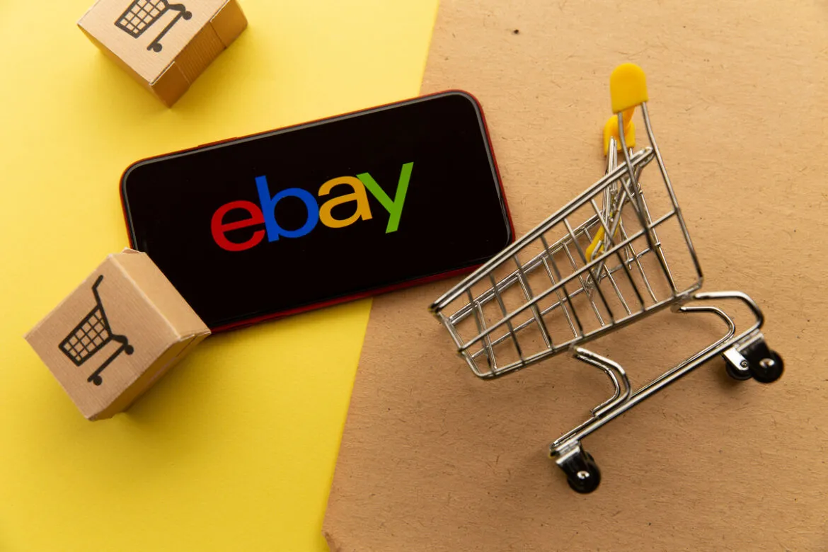 Foto de um celular com o logotipo do Ebay na tela, um carrinho de compras e caixas do lado