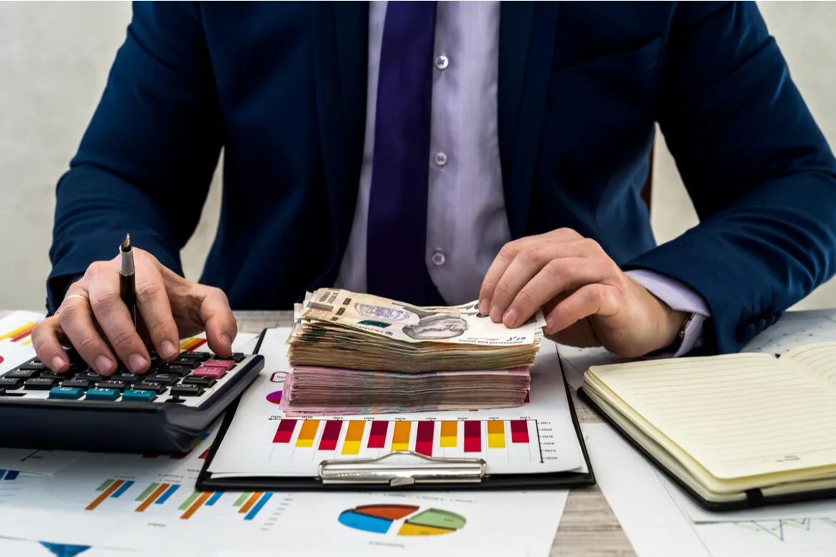 foto de um homem de terno com uma prancheta, calculadora e uma pilha de dinheiro em cima de uma mesa