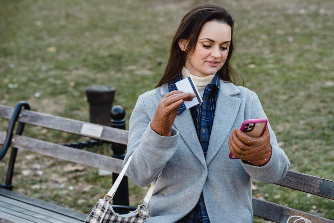 Uma mulher está de pé segurando um celular com a mão direita e com a mão esquerda segurando um cartão de crédito internacional