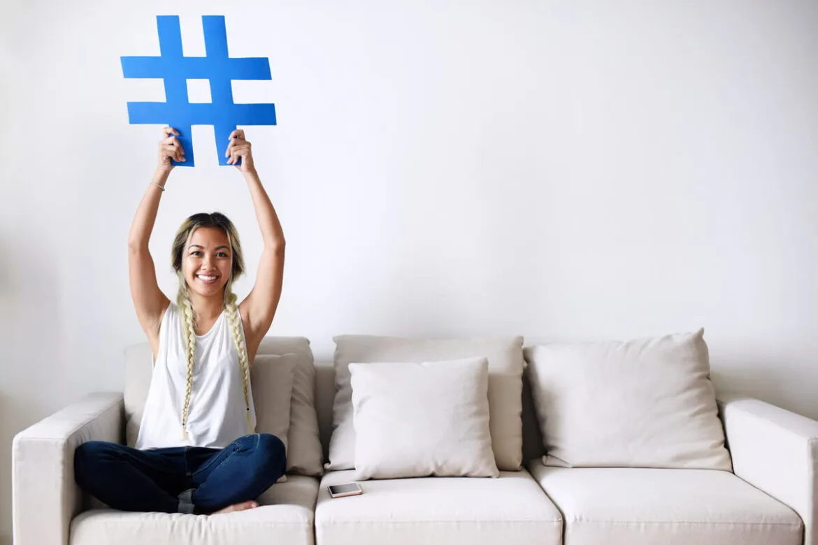 Uma moça sentada em um sofá, segurando uma Hashtag azul com as duas mãos sobre a cabeça