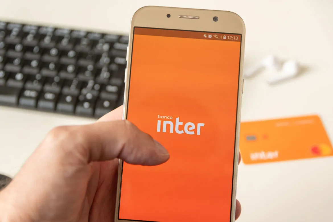 Foto de uma mão segurando um celular que está com a tela do app do Inter aberto, em segundo plano há um teclado de um computador e um cartão do Inter em cima da mesa