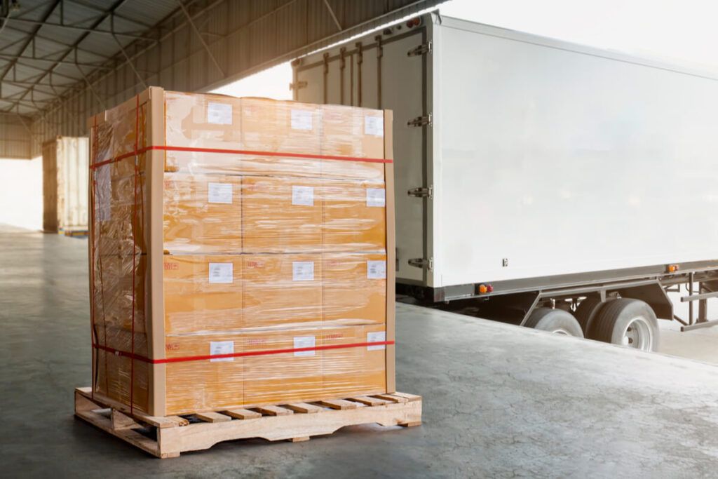 Imagem de um pallet por baixo de uma pilha de caixas em um pátio de transportes e um caminhão parado ao lado
