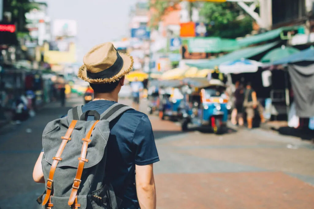 Homem com chapeu e mochila de costas olhando para uma rua com motos e barracas.