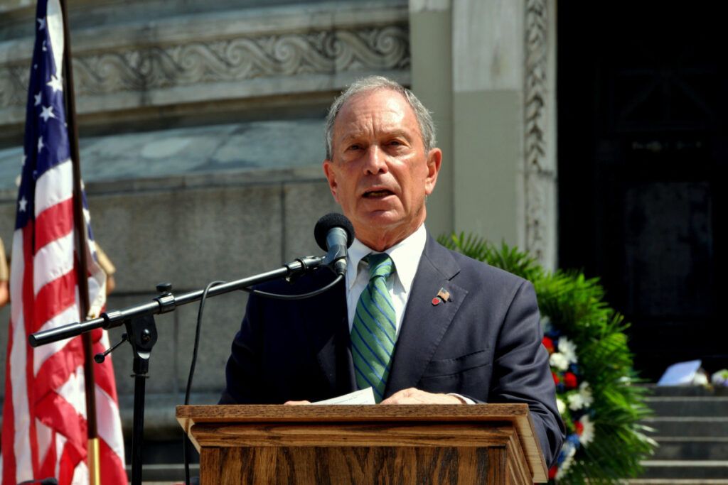 Foto de Michael Bloomberg em frente a um tóten e microfone e ao lado da bandeira dos Estados Unidos